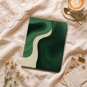 Puede incluir: Un cuaderno verde con un diseño abstracto en capas que presenta tonos de verde y una franja ondulada de color crema. El cuaderno es rectangular y está sobre una superficie suave de color crema. También se ve una taza de café y postales.