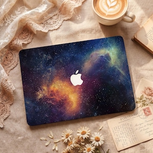 May include: A laptop cover with a colourful nebula design in blues, purples, and oranges, and a white Apple logo. The cover is on a beige surface, with a coffee cup, a book, and a postcard nearby.
