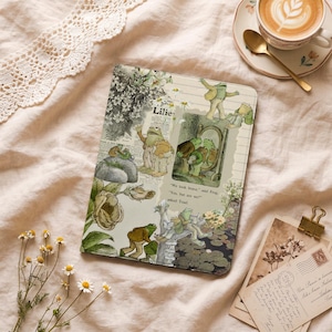 Puede incluir: Un diario decorativo con una ilustración de rana y la palabra "Lilies" en la portada. El diario tiene una estética vintage con elementos florales y botánicos. Una taza de café y una cuchara dorada están en el fondo.