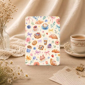 Könnte beinhalten: Eine rechteckige Tablethülle mit einem Aquarellmuster aus Croissants, Eiskaffee, Latte, Beerenkuchen und bunten Schleifen auf cremefarbenem Hintergrund. Das Design enthält kleine florale Akzente und Sternformen, die über die Illustration verteilt sind.