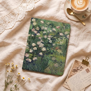 May include: A green notebook with a floral painting of white roses. The notebook is rectangular and lies on a cream-colored fabric. A cup of coffee with latte art and a gold spoon are in the background.