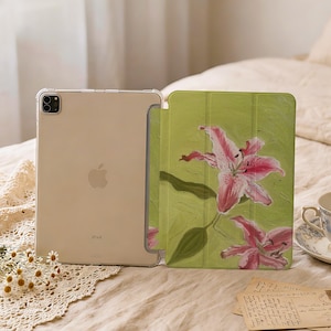 May include: A tablet case featuring a light green background with two pink lily flowers. The case is shown open, revealing a clear back panel that displays the metallic finish and Apple logo of the tablet housed within the protective cover.