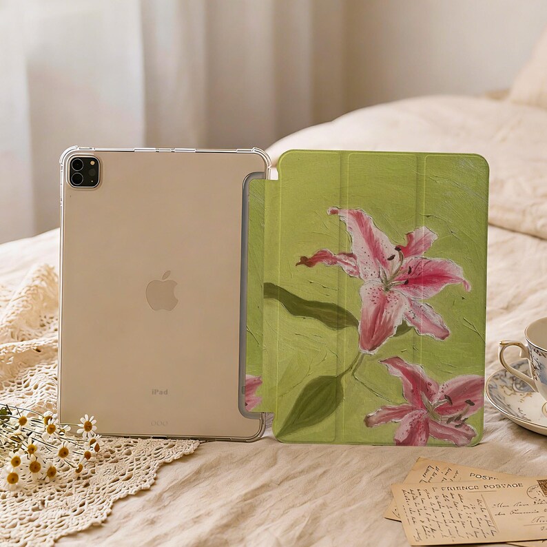 May include: A tablet case featuring a light green background with two pink lily flowers. The case is shown open, revealing a clear back panel that displays the metallic finish and Apple logo of the tablet housed within the protective cover.
