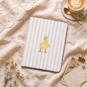 Puede incluir: Un cuaderno con un patrón de rayas rosas y blancas y una ilustración de acuarela de un patito amarillo. El cuaderno está sobre una superficie suave y texturizada, con una taza de café y flores cerca.