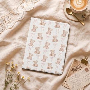 Könnte beinhalten: Eine weiße Tablethülle mit einem sich wiederholenden Muster aus hellbraunen Teddybären und rosa Schleifen. Die Hülle liegt auf einem weichen, neutral getönten Stoff, mit einer Tasse Kaffee und Postkarten im Hintergrund.