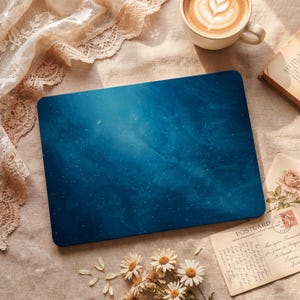 May include: A blue laptop skin featuring a starry night sky design with a crescent moon. The skin is rectangular and sits on a beige surface, surrounded by a cup of coffee, a book, and dried flowers.