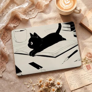 May include: A black cat illustration on a laptop case, lounging on an open book. The case features a black cat with white eyes, a coffee cup, and a pen. A bookmark and a postcard are also visible. The background is a beige surface.