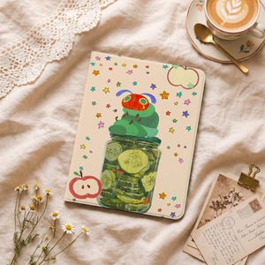 Puede incluir: Una funda rectangular para tableta con fondo crema que presenta una colorida ilustración de una oruga verde con cabeza roja sentada sobre un frasco de vidrio lleno de pepinillos, rodeada de pequeñas estrellas multicolores y un icono de manzana roja.