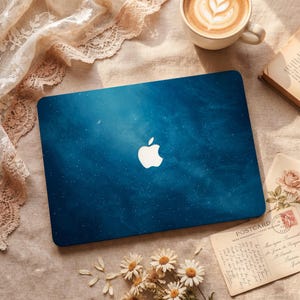 May include: A blue laptop skin with a starry night design and a white Apple logo. The skin features a crescent moon and small white stars. The laptop skin is on a beige surface with a cup of coffee, a book, and a postcard.