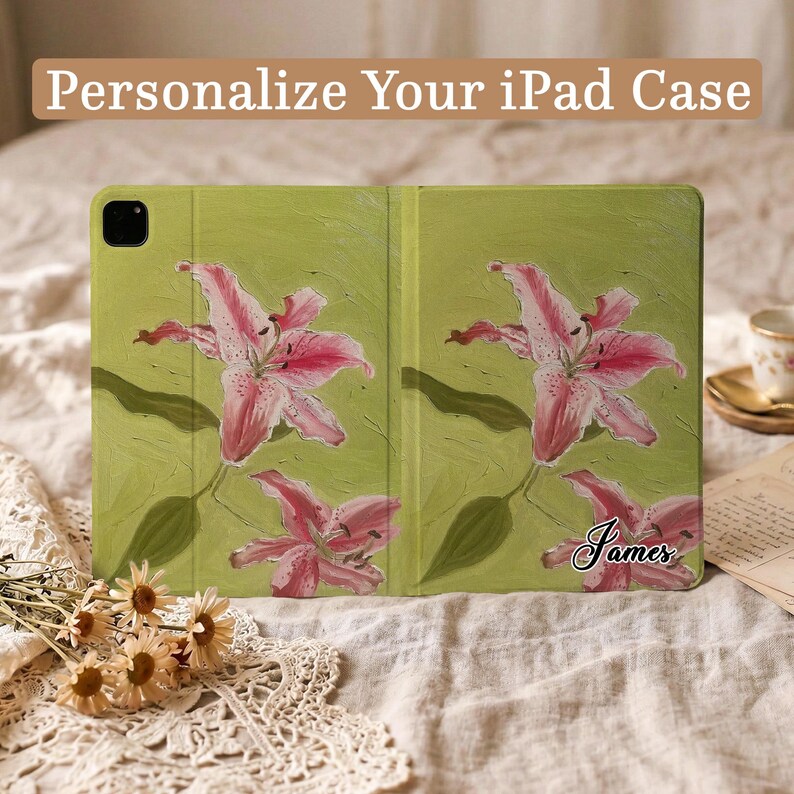 May include: A green iPad case featuring a pink lily floral pattern and the name James printed in black script. The text Personalize Your iPad Case is visible at the top. The case has a camera cutout and a textured, painted appearance.