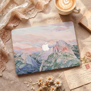 以下が含まれることがあります： 青、ピンク、緑の色合いで山と谷の水彩画の風景が描かれたラップトップケース。 Appleのロゴが見えます。 コーヒーカップ、本、ポストカードも画像にあります。