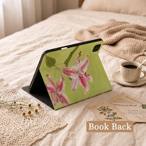May include: A tablet case featuring a painted design of two pink lilies on a light green background. The case is black with a cutout for the camera lens and is positioned in a landscape orientation, displaying the floral artwork on its front cover.