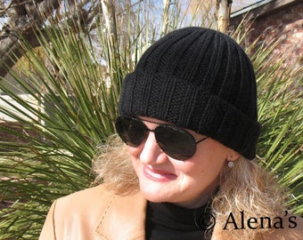 May include: A woman wearing a black knit beanie hat with a brown leather jacket. The hat has a ribbed texture and the jacket has a lapel collar. The woman is wearing sunglasses and has blonde hair.