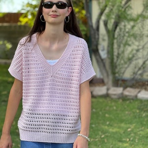 May include: A light pink, short-sleeved, open-weave knit top with a V-neck. The top is paired with a denim skirt. The person is wearing sunglasses and hoop earrings. A white undershirt is visible beneath the knit top. A pearl bracelet is worn on the wrist.