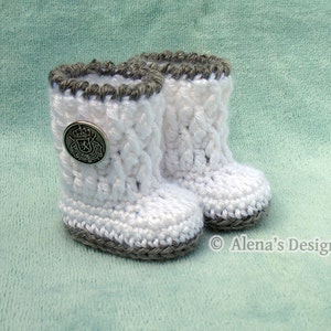 May include: A pair of white crocheted baby booties with gray trim. The booties have a button on the front with a crest design. The booties are on a light green background.