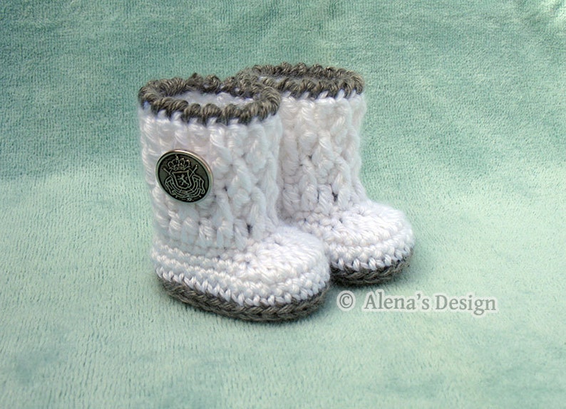 May include: A pair of white crocheted baby booties with gray trim. The booties have a button on the front with a crest design. The booties are on a light green background.
