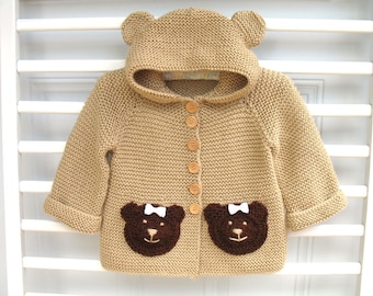 May include: A tan knitted cardigan with a hood shaped like a bear's head. The cardigan has brown bear appliques on the front pockets and wooden buttons.