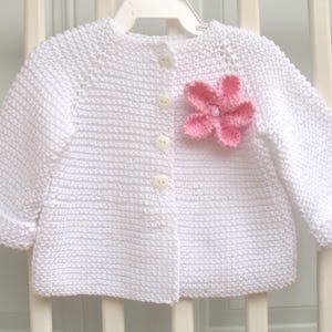 May include: White knitted baby cardigan with a pink flower embellishment. The cardigan has a button closure and long sleeves.