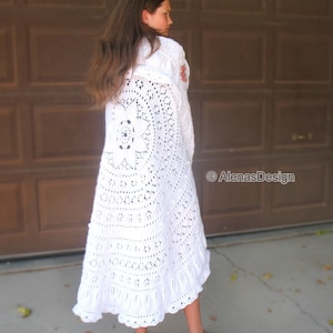 May include: A white crocheted shawl with a large floral design. The shawl is worn by a person standing in front of a brown wooden garage door.
