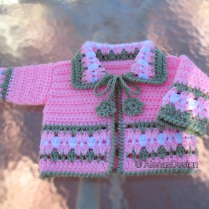 May include: A pink crocheted baby cardigan with a white and green trim. The cardigan has a tie closure and a scalloped edge.