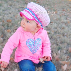 May include: A young child wearing a pink and blue crocheted sailor hat with a heart-shaped design on their pink sweater. The child is sitting on a bed of brown leaves.