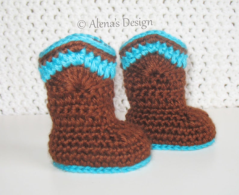 May include: A pair of brown crocheted baby booties with a blue trim. The booties are on a white crocheted surface.