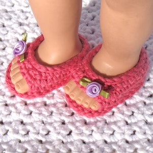 K&ouml;nnte beinhalten: Ein Paar rosa geh&auml;kelte Sandalen mit violetten Blumenverzierungen. Die Sandalen liegen auf einer wei&szlig;en geh&auml;kelten Oberfl&auml;che.