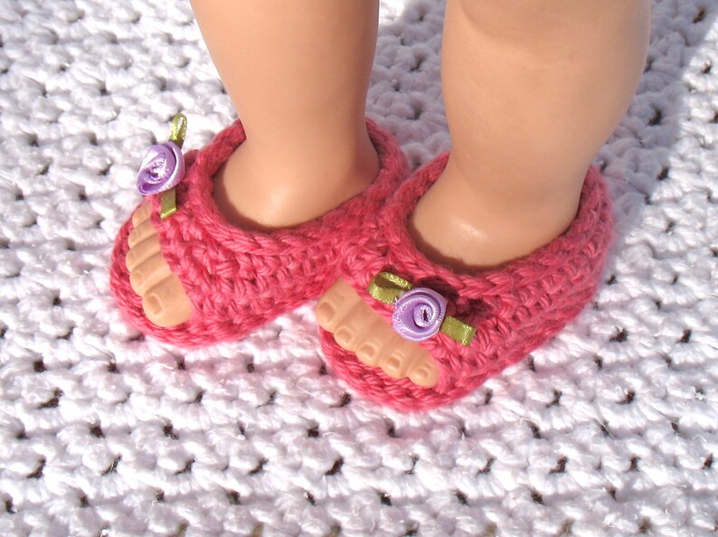 K&ouml;nnte beinhalten: Ein Paar rosa geh&auml;kelte Sandalen mit violetten Blumenverzierungen. Die Sandalen liegen auf einer wei&szlig;en geh&auml;kelten Oberfl&auml;che.