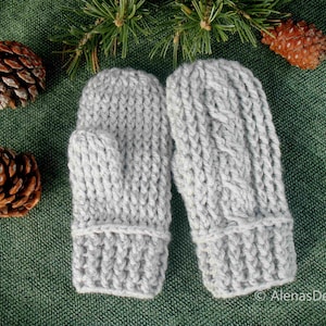 May include: A pair of gray knitted mittens with a cable knit pattern. The mittens are photographed on a green fabric background with pine branches and pine cones.