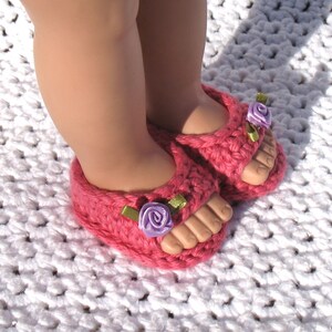 K&ouml;nnte beinhalten: Ein Paar rosa geh&auml;kelte Sandalen mit violetten Blumenverzierungen und gr&uuml;nen Schleifenakzenten.