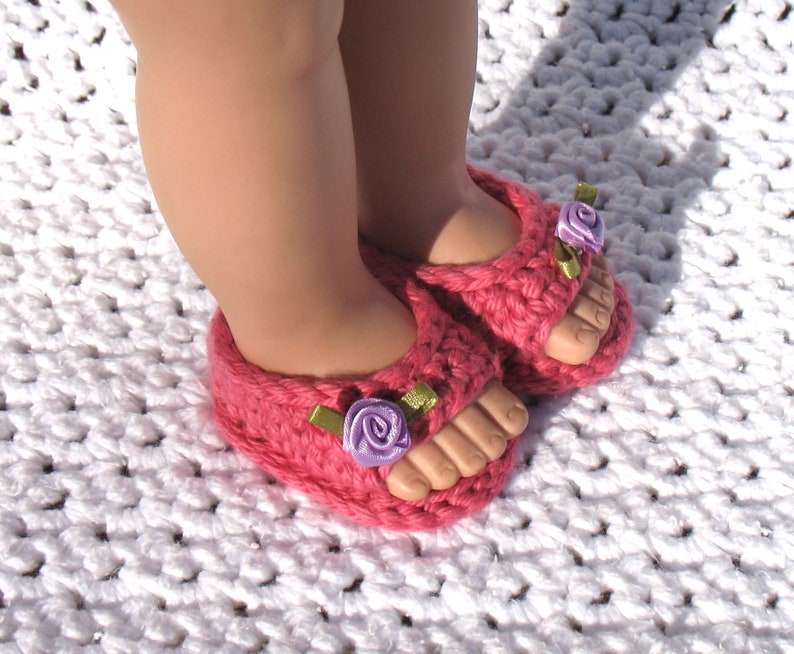 K&ouml;nnte beinhalten: Ein Paar rosa geh&auml;kelte Sandalen mit violetten Blumenverzierungen und gr&uuml;nen Schleifenakzenten.