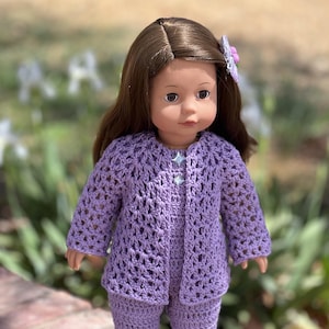 May include: A purple crocheted cardigan and trousers set for a doll. The cardigan has a button fastening and the set features a star-shaped button.