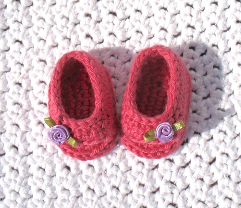K&ouml;nnte beinhalten: Ein Paar rosa geh&auml;kelte Babyschuhe mit lila und gr&uuml;nen Blumenverzierungen.