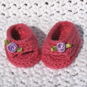 K&ouml;nnte beinhalten: Ein Paar rosa geh&auml;kelte Babyschuhe mit violetten Blumenverzierungen.