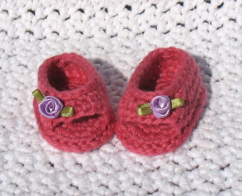 K&ouml;nnte beinhalten: Ein Paar rosa geh&auml;kelte Babyschuhe mit violetten Blumenverzierungen.