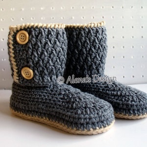 Könnte beinhalten: Ein Paar graue Häkelpantoffeln mit beigefarbenem Besatz und zwei Holzknöpfen an jedem Pantoffel. Die Pantoffeln befinden sich auf einem weißen Hintergrund.