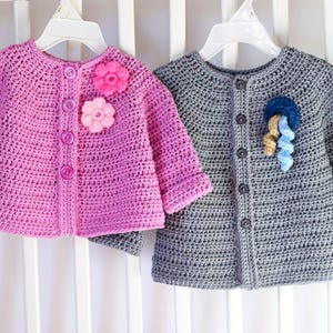 May include: Two crocheted baby cardigans, one pink with pink flower embellishments and one gray with blue and yellow flower embellishments.