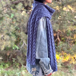 Peut inclure: Une longue écharpe en tricot épais de couleur bleu pervenche profond, présentant un motif de torsades verticales sur toute sa longueur, drapée sur une veste en jean bleu délavé avec un détail d'ourlet effiloché sur le côté inférieur.