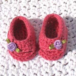 K&ouml;nnte beinhalten: Ein Paar rosa geh&auml;kelte Babyschuhe mit violetten Blumenverzierungen und gr&uuml;nen Schleifenakzenten.