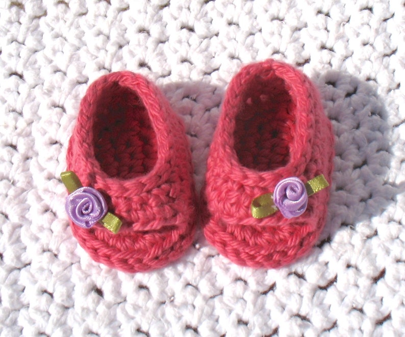 K&ouml;nnte beinhalten: Ein Paar rosa geh&auml;kelte Babyschuhe mit violetten Blumenverzierungen und gr&uuml;nen Schleifenakzenten.