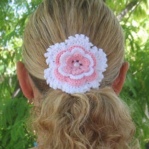 May include: A handmade crochet hair clip in the shape of a flower. The flower has white, pink, and light pink petals, with a pearl bead center. The hair accessory is attached to a blonde ponytail.