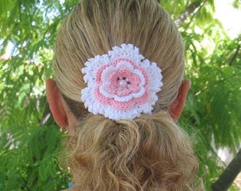 May include: A handmade crochet hair clip in the shape of a flower. The flower has white, pink, and light pink petals, with a pearl bead center. The hair accessory is attached to a blonde ponytail.