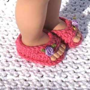 K&ouml;nnte beinhalten: Rosa geh&auml;kelte Sandalen mit violetten Blumenverzierungen. Die Sandalen werden von einem Kind auf einer wei&szlig;en geh&auml;kelten Oberfl&auml;che getragen.