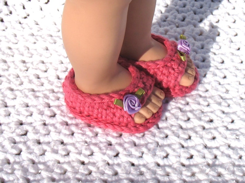 K&ouml;nnte beinhalten: Rosa geh&auml;kelte Sandalen mit violetten Blumenverzierungen. Die Sandalen werden von einem Kind auf einer wei&szlig;en geh&auml;kelten Oberfl&auml;che getragen.