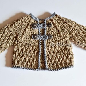 May include: A crocheted baby cardigan in a beige color with gray trim and silver buttons. The cardigan has a textured stitch pattern and a slightly flared shape.