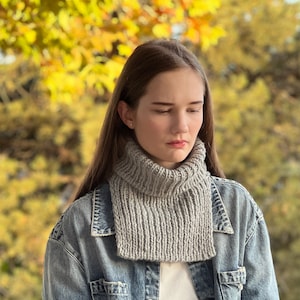 May include: A woman wearing a grey knitted cowl neck scarf over a denim jacket. The scarf is a simple design with a ribbed texture. The woman is standing in front of a blurred background of autumn foliage.
