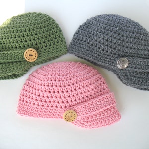 May include: Three crocheted hats in different colors: green, gray, and pink. Each hat has a button on the side.