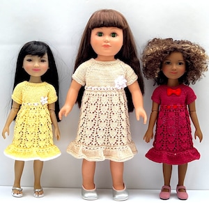 Puede incluir: Tres muñecas con vestidos de punto. La muñeca de la izquierda lleva un vestido amarillo con ribetes blancos. La muñeca del centro lleva un vestido beige con ribetes blancos. La muñeca de la derecha lleva un vestido rosa con un lazo rojo.