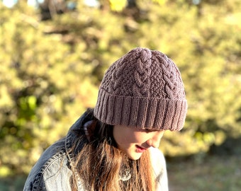 May include: A mauve knitted beanie with a cable knit pattern. The beanie is worn by a person with long brown hair.