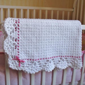 May include: White crocheted baby blanket with a pink crocheted border and a pink satin ribbon bow.
